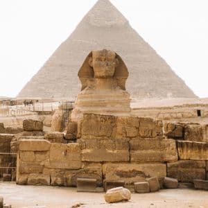 Un'imponente statua antica di sfinge si erge di fronte a una massiccia piramide di pietra, circondata da rovine su terreno sabbioso.