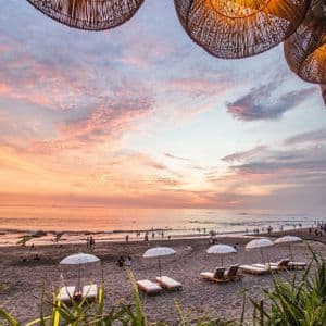Paralumi intrecciati pendono sopra una spiaggia sabbiosa con lettini e ombrelloni mentre le persone si godono un tramonto colorato sull'oceano.