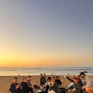 Un grupo de viaje de WeRoad se sienta en mantas en una playa de arena, disfrutando de bebidas durante un atardecer dorado sobre el océano.