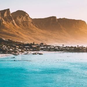 Ein Panoramablick auf eine Küstenstadt mit einem weißen Sandstrand und türkisfarbenem Wasser am Fuße einer zerklüfteten Bergkette bei Sonnenaufgang.