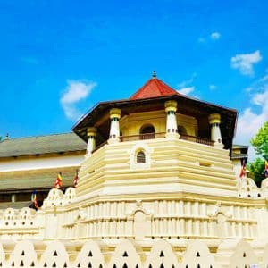 Un tempio bianco a più livelli con una torre ottagonale e tetto rosso si erge dietro un muro decorativo sotto un cielo blu.