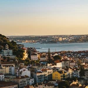 Un paesaggio urbano con edifici colorati e tetti rossi si affaccia su un ampio fiume al tramonto, con un castello storico su una collina.
