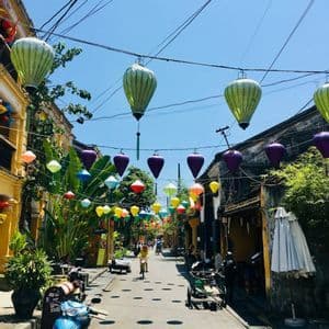 Lanterne colorate adornano una strada stretta e illuminata dal sole, fiancheggiata da edifici gialli e scooter parcheggiati.