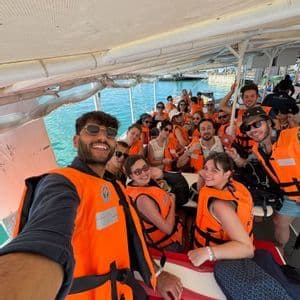 A WeRoad group trip takes a selfie on a boat, all wearing orange life jackets over turquoise water.
