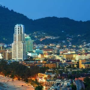 An illuminated coastal city with modern buildings nestled between a sandy beach and lush green hills at twilight.