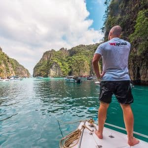 A man from a WeRoad trip stands on the bow of a boat, looking out over a turquoise bay with rocky cliffs.