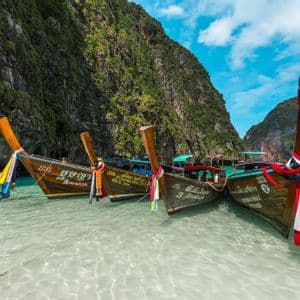 A WeRoad group trip swims in a clear lagoon next to traditional long-tail boats moored by steep green cliffs.