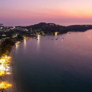An aerial view of a brightly lit coastal town and beach next to a calm bay with boats at dusk.