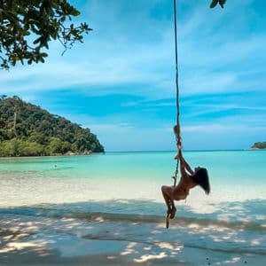 A woman in a bikini swings on a rope over the turquoise sea from a white sandy beach with lush hills in the background.
