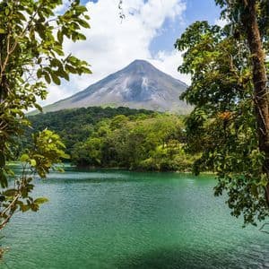 A large volcano rises above a dense forest and a green lake, framed by leafy trees under a partly cloudy sky.