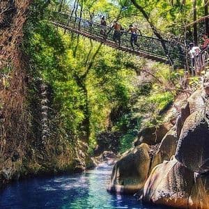 Un viaggio di gruppo WeRoad attraversa un ponte sospeso su un fiume turchese che scorre attraverso un canyon roccioso e alberato.