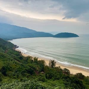 A high-angle view of a tropical coastline with a sandy bay, lush green hills, and distant mountains under an overcast sky.