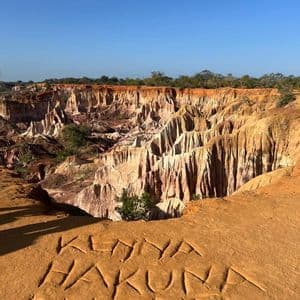 The words 'Kenya Hakuna Matata' carved into the orange sand on a cliff's edge, overlooking a vast, multi-colored canyon.