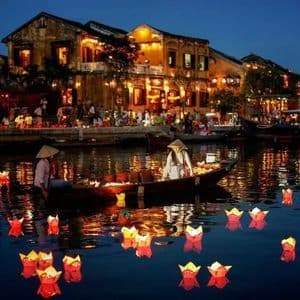 Due persone in una barca tradizionale circondate da lanterne di carta galleggianti su un fiume di notte, con edifici illuminati sullo sfondo.