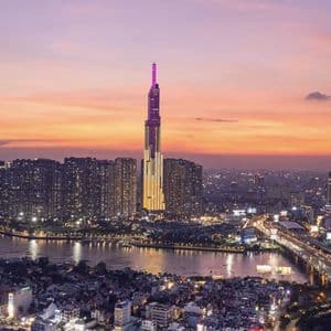 Un grattacielo illuminato con una guglia viola domina un denso skyline cittadino accanto a un ampio fiume al tramonto.