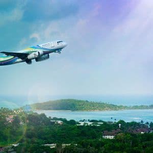 Un colorato aereo passeggeri vola basso su una lussureggiante costa tropicale con verdi colline e piccole isole nel mare.