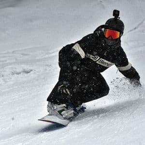 Uno snowboarder con casco e telecamera scende un pendio innevato, sollevando neve fresca mentre nevica.