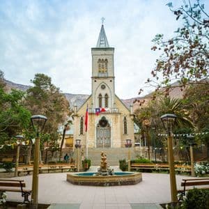 Una chiesa in pietra con un alto campanile e due bandiere si erge in una piazza con una fontana, circondata da alberi e montagne.