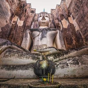 Eine kolossale, verwitterte Stein-Buddha-Statue sitzt meditierend zwischen hohen Steinmauern, mit einer kleinen Opferschale davor.