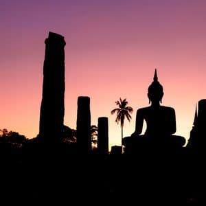 Siluetas de una estatua de Buda sentado, ruinas antiguas y una palmera contra un atardecer morado y naranja.