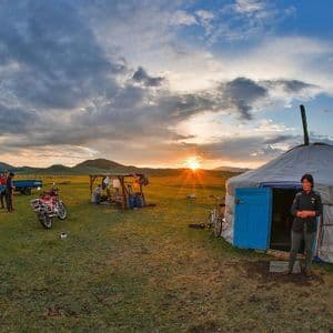 Bei Sonnenuntergang steht eine Person vor einer traditionellen Jurte auf einem Campingplatz auf einer grasbewachsenen Ebene, im Hintergrund Fahrzeuge und Berge.