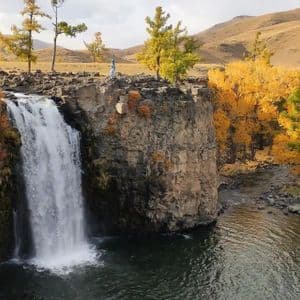 Eine Person in Blau steht mit erhobenen Armen auf einer felsigen Klippe und blickt auf einen Wasserfall, umgeben von Herbstbäumen.
