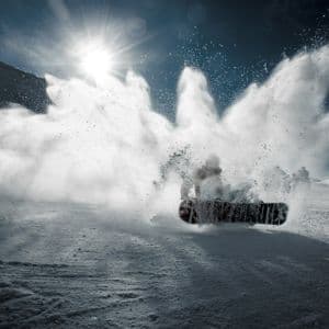 A snowboarder carves through fresh powder on a mountain slope, creating a large spray of snow under the bright sun.