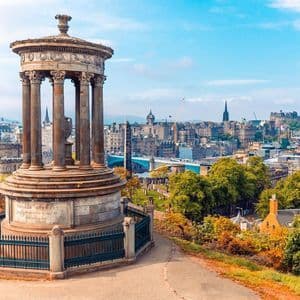 Ein klassisches Steindenkmal auf einem Hügel, das eine historische Stadtlandschaft voller herbstlich gefärbter Bäume unter blauem Himmel überblickt.