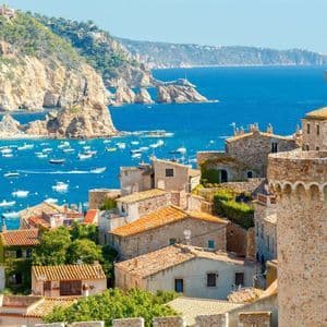 Una cittadina costiera, con edifici in pietra e una torre, si affaccia su una baia azzurra, piena di piccole imbarcazioni bianche e una spiaggia a ridosso della scogliera.