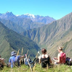 A WeRoad group trip rests on a grassy mountainside, looking out at a vast valley and distant snow-capped peaks.