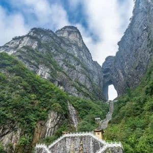 Una larga escalera asciende por una montaña empinada y cubierta de bosque hacia un gran arco de roca natural bajo un cielo nublado.