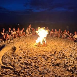 A WeRoad group trip gathered around a bonfire on a beach at night, with a large dolphin drawn in the sand in the foreground.