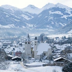 Blick von oben auf ein schneebedecktes Alpendorf mit einer markanten Kirche vor schneebedeckten Bergen.