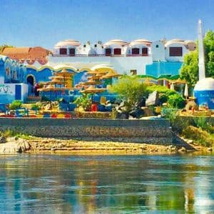 Colorati edifici blu e bianchi in stile nubiano su una sponda rocciosa del fiume, visti dall'acqua.