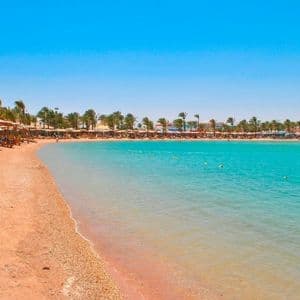 Una spiaggia di sabbia curva incontra acque calme e turchesi, con palme e ombrelloni di paglia che costeggiano la riva sotto un cielo limpido.