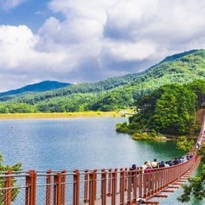 Persone che passeggiano su un lungo ponte sospeso sopra un lago, con montagne verdi e boscose sullo sfondo.