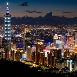 Una vista panoramica di uno skyline cittadino esteso e illuminato di notte, con un alto e distintivo grattacielo che si erge sopra gli altri edifici.