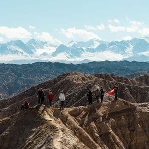 A WeRoad group trip stands on a ridge overlooking a vast canyon landscape, with a distant snow-capped mountain range under a blue sky.