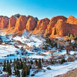 Sunlight illuminates large red rock formations towering above a snow-covered valley dotted with pine trees and a small village.