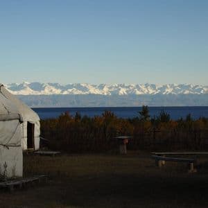 Two yurts on the shore of a large lake, with a distant snow-capped mountain range visible under a clear blue sky.