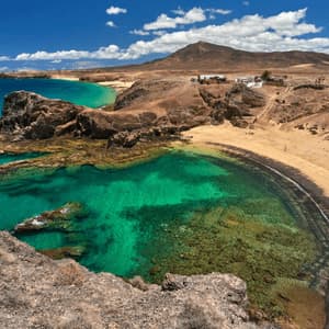 Una vista aérea de una cala apartada con agua turquesa cristalina, una playa de arena y un paisaje rocoso bajo un cielo azul.