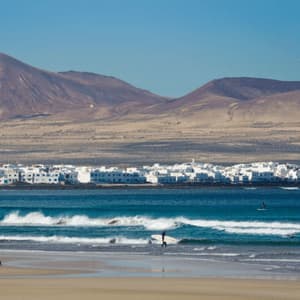 Una playa de arena con gente y surfistas en el océano, con un pueblo costero blanco y montañas áridas al fondo.