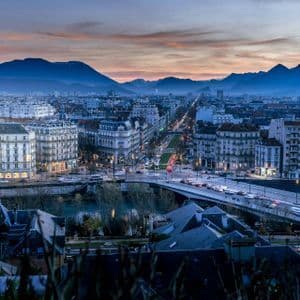 Una veduta dall'alto di una città al tramonto, con strade ed edifici illuminati e una catena montuosa sullo sfondo.