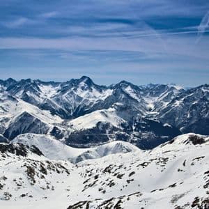 Una vista panoramica di una vasta catena montuosa innevata sotto un cielo azzurro con nuvole sottili e scie di condensazione.
