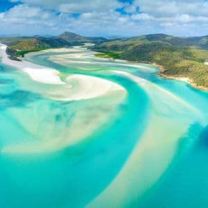 An aerial view of swirling white sandbars in vibrant turquoise water, with green, hilly islands in the background under a blue sky.