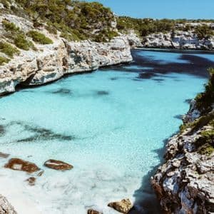 Una vista da un'alta scogliera di una caletta appartata con acqua turchese trasparente, una piccola spiaggia di sabbia bianca e coste rocciose.