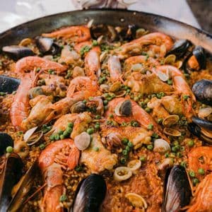 Primo piano di una paella tradizionale in una grande padella, ricca di riso, gamberi, cozze, pollo e piselli.