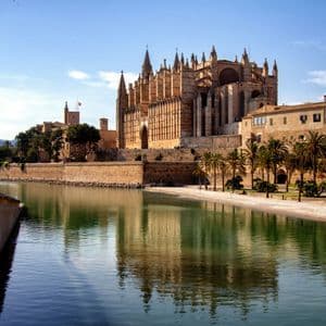 Una gran catedral gótica y edificios adyacentes reflejados en las aguas tranquilas de un lago, con palmeras bordeando la orilla.