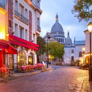 Una strada acciottolata con caffè dalle luci calde e una grande basilica dalla cupola bianca sullo sfondo al tramonto.