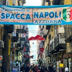 Un grande striscione blu per 'Spaccanapoli Azzurra' pende su una strada stretta e affollata fiancheggiata da negozi ed edifici gialli.
